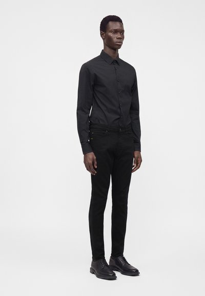 Modello maschile in piedi che indossa una camicia nera con bottoni, pantaloni neri e scarpe eleganti nere su uno sfondo bianco semplice.