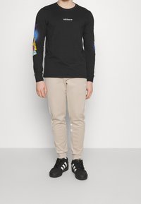 Mężczyzna ubrany w czarną koszulę Adidas z długim rękawem z kolorowymi grafikami na rękawach, beżowe spodnie dresowe Adidas oraz czarne sneakersy Adidas z białymi paskami.