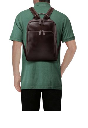 Homme portant un sac à dos en cuir marron foncé avec deux compartiments zippés, une chemise à manches courtes verte et un pantalon noir, vu de dos.