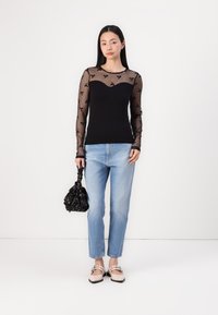 Schwarzes Langarmoberteil mit durchsichtiger gepunkteter Netzstruktur und floralen Akzenten, kombiniert mit hellblauen Jeans in gerader Form und flachen Schuhen. Hält eine schwarze Tasche.