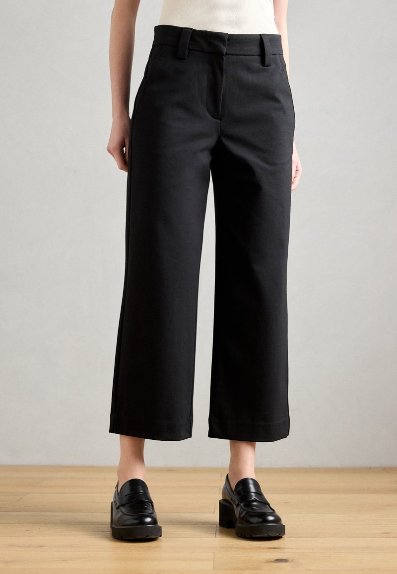 Sorte bukser med vide ben lavet af et glat stof med høj talje, bæltestropper og en ren finish nederst. Kombineret med sorte loafers.