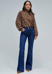 Blusa de estampado de serpiente marrón con cuello alto y detalle de lazo, combinada con jeans de campana de color azul oscuro y tacones puntiagudos de color beige.