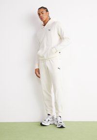Sudadera blanca y joggers a juego hechos de un tejido suave, con un logo negro en el pecho y la pierna del pantalón. Las zapatillas tienen un diseño en blanco y negro.