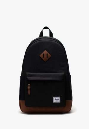 Schwarzer Rucksack mit braunem Boden und Lederpatch, vordere Tasche mit Reißverschluss und Stoffmarkenetikett in der unteren rechten Ecke der Tasche.