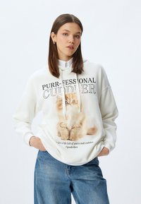 Witte hoodie met een afbeelding van een pluizige kat en de tekst "PURR-FESSIONAL CUDDLER." Van katoen, met een losse pasvorm en een buidelzak.
