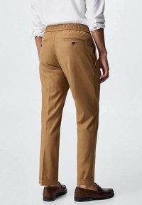 Mann trägt taillierte, camel-farbene Hose mit elastischem Bund, kombiniert mit braunen Slippern und einem weißen Hemd mit hochgekrempelten Ärmeln.