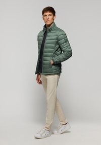 Chaqueta acolchada verde con un alto cuello, cierre de cremallera y bolsillos laterales. Llevada sobre una camisa negra, combinada con pantalones beige claro y zapatillas blancas.