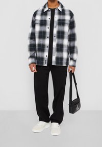 Camicia di flanella a quadri bianca e nera con chiusura a bottoni, due tasche sul petto, indossata sopra una t-shirt nera, abbinata a pantaloni neri e sneakers bianche.