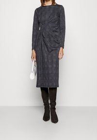 Robe à manches longues grise à carreaux avec un design à l'avant torsadé, associée à des bottes noires en suédine montant jusqu'au genou et un sac à main blanc.