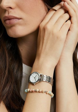 Silberne Metall-Armbanduhr mit weißem Zifferblatt und Gliederarmband, kombiniert mit einem mehrfarbigen Perlenarmband, das an einem Handgelenk getragen wird.
