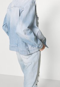 Giacca in denim blu chiaro con vestibilità rilassata, dotata di chiusura a bottoni, tasche sul petto e dettagli leggermente usurati ai bordi.