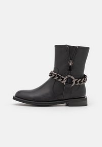 Bota negra hasta el tobillo hecha de cuero suave con punta redondeada, cremallera lateral y un detalle de cadena plateada alrededor del tobillo. Tacón bajo en bloque.