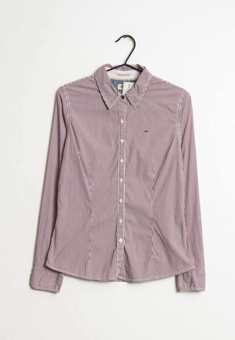 Chemise à manches longues boutonnée avec fines rayures verticales rouges et blanches suspendue sur un cintre noir contre un fond blanc.