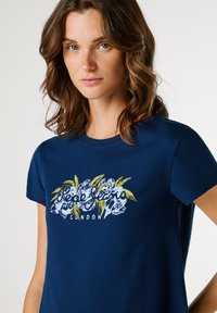 Μπλε βαμβακερό t-shirt με λουλουδάτο γραφικό τύπωμα σε λευκό και πράσινο, με το κείμενο "Pepe Jeans London" στο κέντρο κάτω από το σχέδιο.