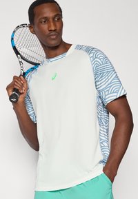 Λευκό αθλητικό μπλουζάκι με κοντά μανίκια και μπλε μοτίβο κυμάτων στους ώμους, με πράσινο λογότυπο, φορεμένο από άτομο που κρατά ρακέτα τένις.
