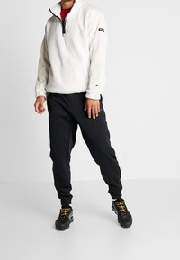 Osoba ubrana w białą polarową bluzę z zamkiem do 1/4, czarne dresowe spodnie oraz czarne sneakersy z żółtymi sznurówkami, stojąca na tle jednolitego białego tła.