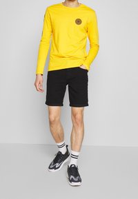 Camisa amarilla de manga larga con un logo espiral negro en el pecho, combinada con pantalones cortos negros. Zapatillas negras y blancas completan el look.