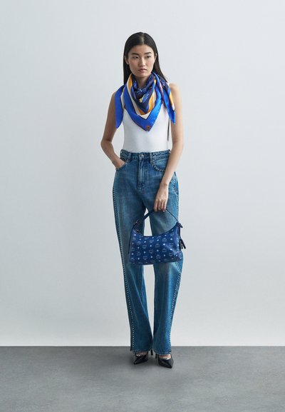Frau trägt ein weißes, ärmelloses Top, blaue Jeans mit Seitenstiften, einen blauen gemusterten Schal, schwarze High Heels und hält eine blaue Monogramm-Tasche vor einer neutralen Wand.