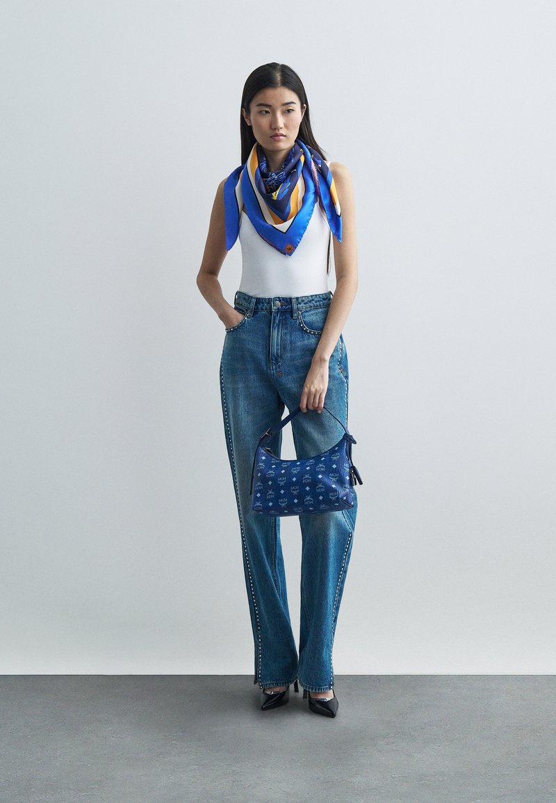 Kvinde iført hvid ærmeløs top, blå jeans med side pynteknapper, blå mønstret tørklæde, sorte hæle, holder en blå monogram håndtaske op ad en neutral væg.