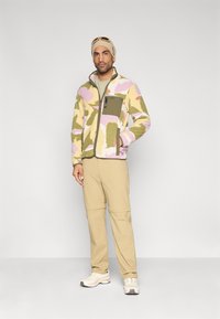 Ζακέτα fleece σε ροζ, πράσινο και μπεζ καμουφλάζ μοτίβο με τσεπάκι με φερμουάρ. Μπεζ παντελόνι και αθλητικά παπούτσια ολοκληρώνουν το σύνολο.