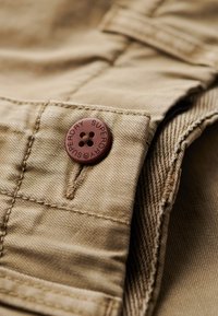 Tan stof met een geweven structuur, voorzien van een bruine knop die is gegraveerd met "SUPERDRY" en een viergaatjes sluitdetail.