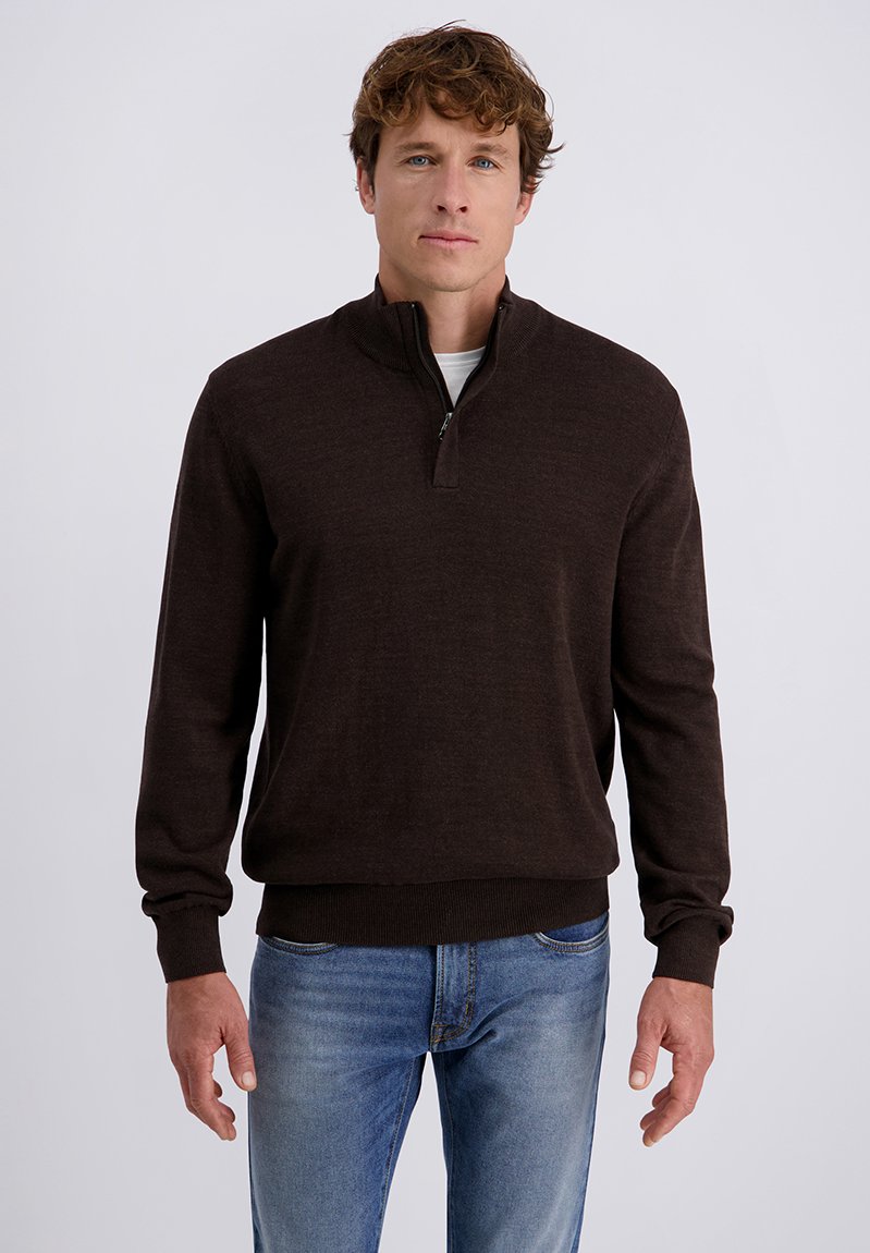 Uomo con capelli ricci castani che indossa un maglione marrone scuro con zip fino al quarto e jeans blu, in piedi contro uno sfondo chiaro e semplice.