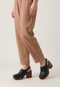 Person trägt hellbraune Leinenhose und schwarze Leder-Clogs mit klobigen Holzabsätzen und Knöchelriemen vor hellem Hintergrund.