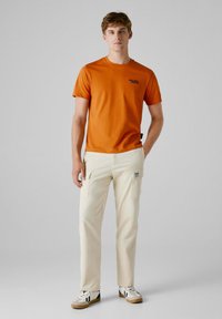 Orange bomulds t-shirt med et lille sort logo, parret med krèm cargo bukser, der har sidelommer og en lappem, samt hvide sko med sorte detaljer.