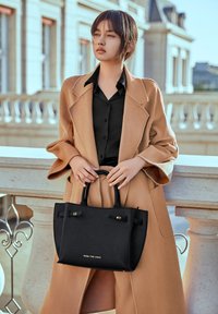 Zwarte leren handtas met een gestructureerd oppervlak, korte handvatten en gouden hardware accenten. Gecombineerd met een beige oversized jas en een zwart overhemd.