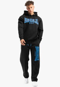 Črni komplet z kapuco in trenirkami z logotipom Lonsdale v modri barvi. Tkanina se zdi mehka, s sproščenim krojem in zankami za prilagajanje.