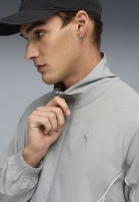 Lys grå zip-up jakke med høj krave, elastiske manchetter og et logo på venstre bryst. Har en glat overflade og reflekterende detaljer.