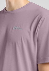 T-shirt a maniche corte di colore lavanda, realizzato in tessuto morbido, con un logo discreto in azzurro chiaro sulla parte sinistra del petto. Texture liscia, design casual.