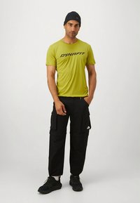 Zelena sportska majica s logotipom "DYNAFIT", crne terenske hlače s džepovima, crne tenisice i tamna kapa. Jednostavan dizajn, funkcionalna odjeća.