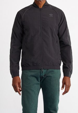 Chaqueta negra con cierre frontal, dobladillo acanalado, bolsillos laterales y tres rayas grises en las mangas. Fabricada en un tejido ligero y texturizado.