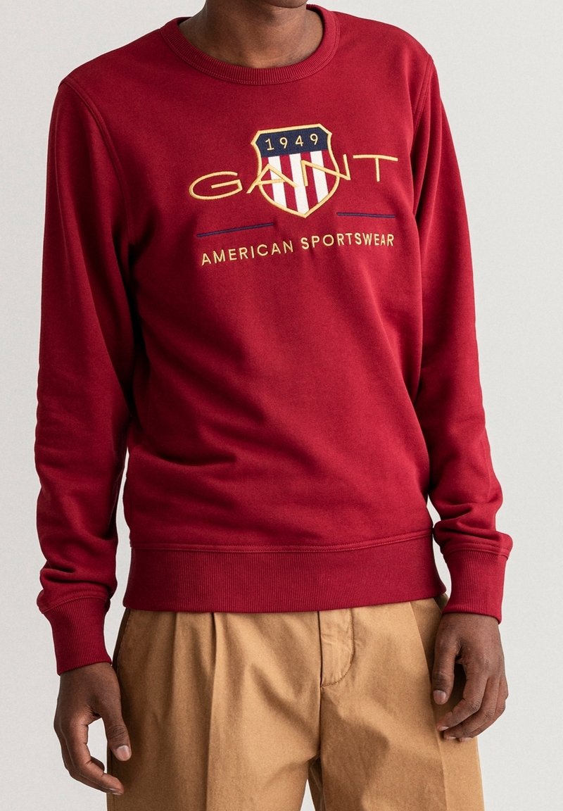 Homme portant un sweat-shirt rouge GANT avec les logos "1949" et "American Sportswear", associé à un pantalon beige, sur un fond uni.