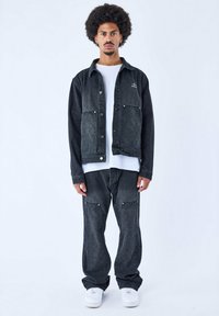 Chaqueta de mezclilla negra con bolsillos frontales acolchados, combinada con pantalones de mezclilla negra a juego, llevada sobre una camiseta blanca. Presenta costuras en contraste.