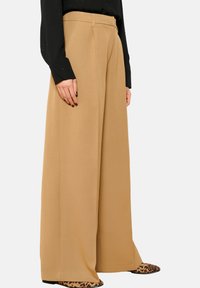 Camelkleurige pantalon met wijde pijpen en plooien, gemaakt van een zachte stof. Gecombineerd met een zwart shirt met lange mouwen en schoenen met luipaardprint.