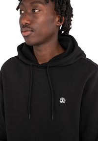 Felpa nera con cappuccio in tessuto morbido, dotata di un cordino per regolare il cappuccio e un piccolo logo bianco sul petto. Design semplice e funzionale.