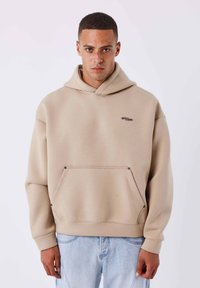 Sudadera con capucha beige hecha de tela suave, que presenta un gran bolsillo frontal, detalles mínimos de costuras y un pequeño logo en el pecho.
