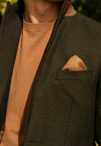 Marynarka w jodełkę w ciemnozielonym kolorze z brązową, teksturowaną poszetką, noszona na jasnobrązowym swetrze. Wyraźnie podkreślone detale.