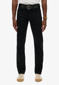 Jean en denim noir avec une coupe slim, design à cinq poches, accompagné d'une ceinture noire et de baskets de couleur claire.