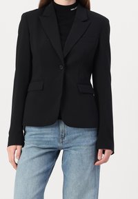 Černý propracovaný blazer s jedním knoflíkem, klopovým límcem a předními kapsami, nošený přes černý top a doplněný světlými modrými džíny.
