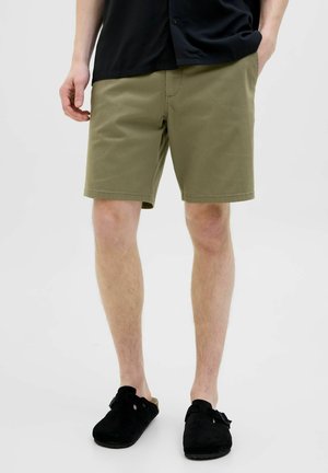 Bărbat care poartă pantaloni scurți verde măsliniu până la genunchi, cămașă neagră și pantofi slip-on negri, stând cu mâinile în buzunare.