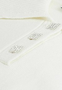 Weißes Top mit Kragen und gerippter Textur, mit drei diamantförmigen Kristallakzent-Knöpfen auf dem Blendenbereich.