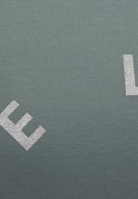 Tessuto verde a coste con dettagli testuali glitterati argentati. La texture è liscia con linee verticali, creando un sottile motivo visivo.