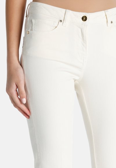 Tuva personas bilde, kas valkā baltas augstu jostasvietu džinsa bikses ar priekšējām kabatām un zelta pogu, roka atrodas tuvumā augšstilbam.