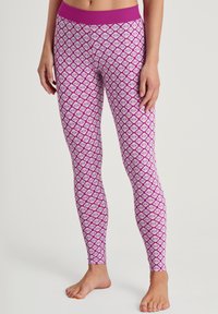 Vrouw in strakke roze en witte gemusterde leggings die blootvoets tegen een effen achtergrond staat.