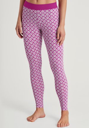 Eine Frau in enganliegenden, pink-weißen gemusterten Leggings steht barfuß vor einem schlichten Hintergrund.