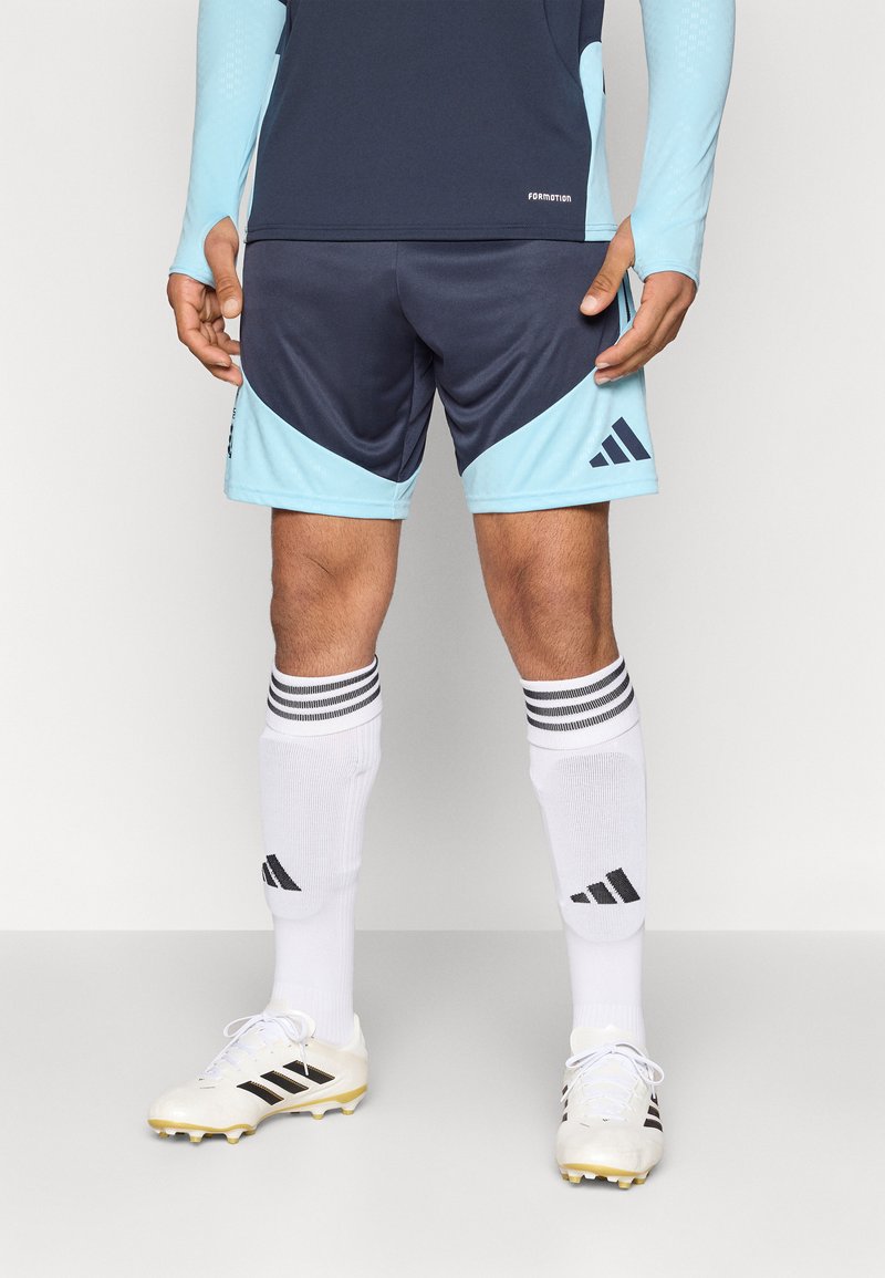 Αθλητική στολή Adidas με ναυτικά σορτς με ανοιχτό μπλε λεπτομέρειες, λευκές κάλτσες με μαύρες ρίγες και λευκά παπούτσια με μαύρες λεπτομέρειες.