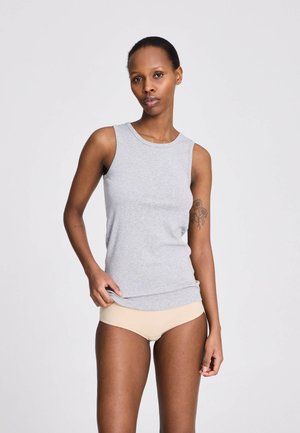 Femeie cu păr scurt stă lângă un fundal simplu, purtând un top gri fără mâneci și lenjerie bej, arătând un tatuaj floral pe braț.